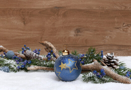Weihnachtlicher Hintergrund im Schnee mit einer blauen Christbaumkugel, Tannenzweigen und Beeren vor einem braunen Holzhintergrund mit viel Platz f&uuml;r Text und eigene Weihnachtsgr&uuml;&szlig;e.