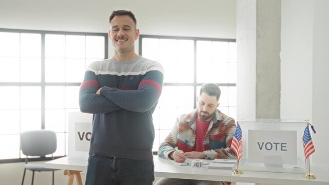 Man stands with arms crossed beside a ballot box and ballot sign in a polling building, smiling while a poll worker fills forms at a desk; civic pride.