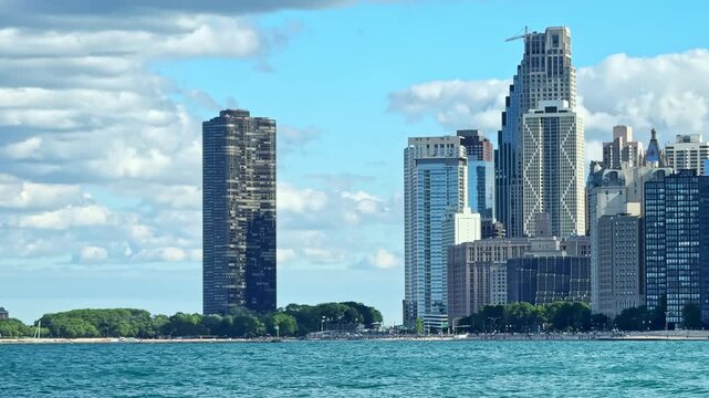 The view shows tall buildings next to a lake in Chicago. The water reflects the city skyline. Clouds are scattered in the blue sky above the scene.