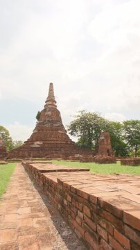 Camera glides along a low brick wall towards the ancient bell-shaped stupa at Wat Chakkrawat in Ayutthaya, Thailand. The historic brick monument sits on a green lawn under a cloudy summer sky.