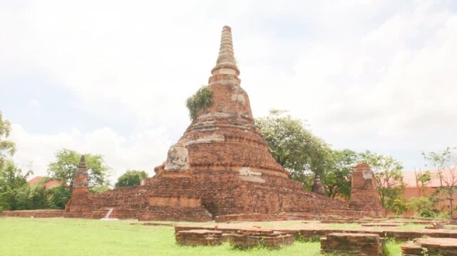 Camera rises over a weathered red brick wall to reveal the ancient brick stupa at Wat Chakkrawat in Ayutthaya, Thailand. The historic monument sits on a green grassy lawn under a cloudy summer sky.