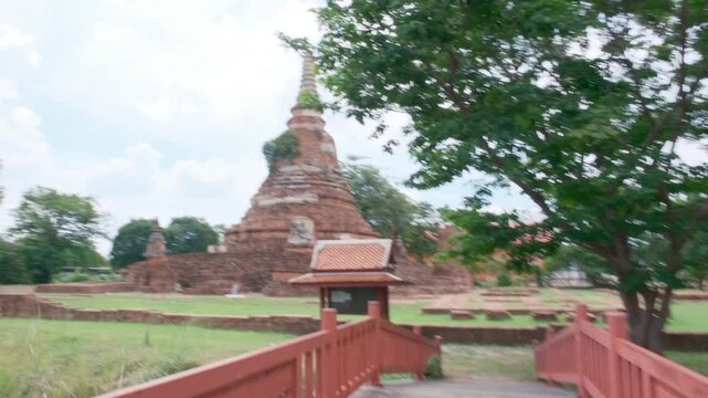 A wooden bridge with red railings leads toward the ancient brick stupa of Wat Chakkrawat in Ayutthaya, Thailand. A small traditional information pavilion stands near green trees under a cloudy sky.