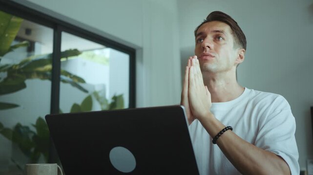 Stressed millennial man at laptop holding hands together and looking up hoping for success business pressure decision moment remote work entrepreneur facing uncertainty home office. Business man prays