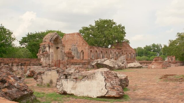 Wide exterior view of the ancient ruins at Wat Kudi Dao in Ayutthaya, Thailand. Large fallen stone debris rests on the grass in front of weathered red brick walls and arched doorways under cloudy sky.