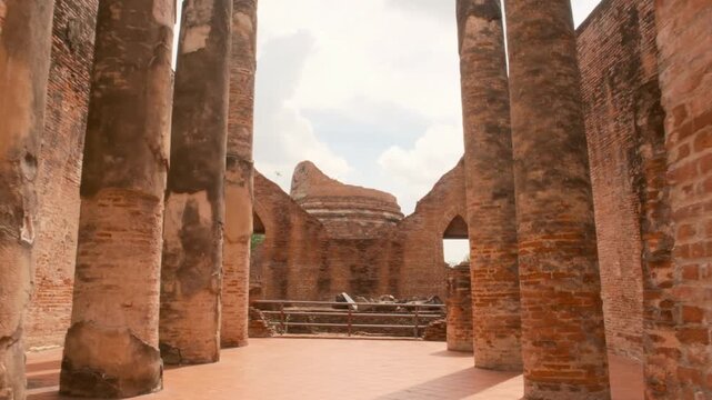 Camera moves slowly through an ancient brick entrance into the ruined assembly hall of Wat Kudi Dao, Ayutthaya, Thailand. Tall pillars and red brick walls frame a broken stupa under a cloudy sky.