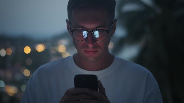 Smartphone addiction concept. Close up young man using smartphone at night outdoors with glowing screen reflected in glasses, symbolizing smartphone addiction. modern lifestyle technology habits.