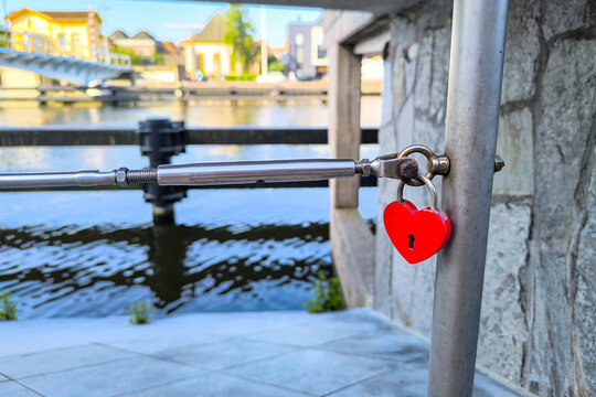 Vivid red heart-shaped padlock fastened to a steel cable railing beneath a bridge overlooking a Dutch canal, with waterside houses and a footbridge softly blurred in the background.