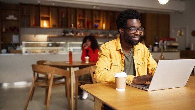 Focused African American man with glasses working on laptop in cafe and drinking hot drink. Male sitting at wooden table with coffee cup, woman blurred in background, modern coffee shop interior.