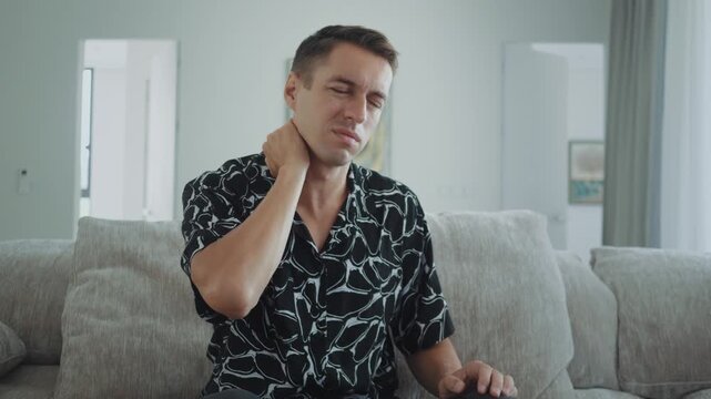 Neck pain concept. Young man sitting on sofa suffering neck pain and hold stiff neck with hand in living room at home showing muscle tension, neck pain problem from sedentary lifestyle and office work