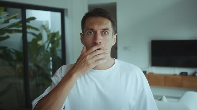 Man in modern apartment stares at camera in shock, removes glasses in fear, eyes wide and mouth tense, reacting to alarming news or sudden threat, expressing panic, anxiety, intense emotional surprise