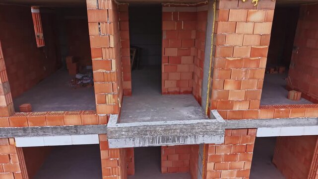 Workers are constructing a building using red bricks. The site is active with materials and tools visible. The structure has open spaces and scaffolding for support.