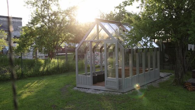 Sunlit wooden greenhouse with tempered glass sits on gravel base, doors open to pots and raised beds. Warm highlights, slight lens flare, gentle branch movement.