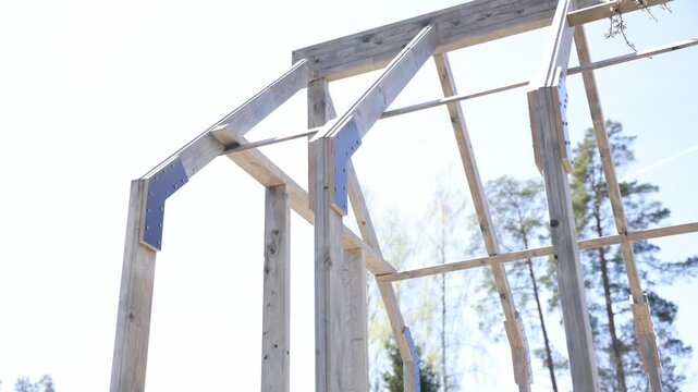 Close up unfinished wooden frame with angled rafters and metal plates. Builder assembles for tempered glass. Camera pans upward in strong sunlight.