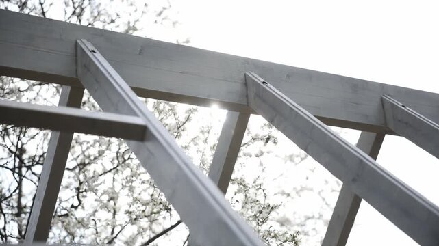 Close up shows painted timber rafters and vertical supports forming a grid. Sun flare filters through tempered glass during a slow upward pan in mid morning.