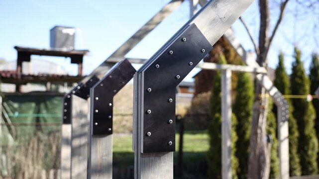 Close up shows wooden posts joined by black connector plates with bolts, forming rafters in a residential garden near a wooden pergola and columnar conifers in daylight.