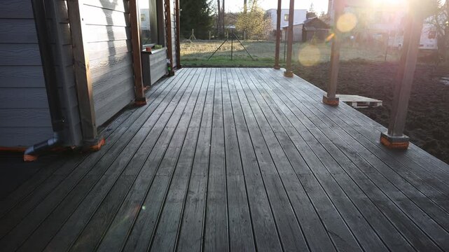 Wooden deck extends from house wall toward yard. Grooved planks, metal posts on orange pads, wet surfaces, lens flare, long shadows, white van, pallet, outbuilding.