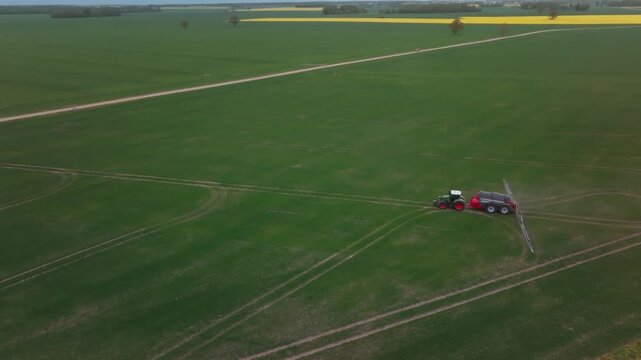 Aerial view shows tractor towing multi wheel sprayer across green crop plots, fine mist spraying, curved turns and parallel tracks by dirt road and yellow flowering strip