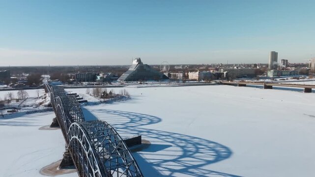 Aerial view shows Riga railway bridge over the frozen Daugava River. Drone moves along the bridge axis as tiny figures cross. National Library and Ferris wheel appear.