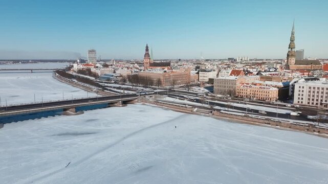 Aerial pans show frozen Daugava River, Old Town roofs, St. Peter's Church and Riga Cathedral. Tram and cars cross the bridge as faint pedestrian tracks mark the ice.