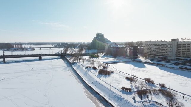 Aerial view of frozen Daugava in Riga with people walking, ski tracks, bridges, National Library of Latvia, Radisson sign, long shadows, lens flare, urban winter scene.