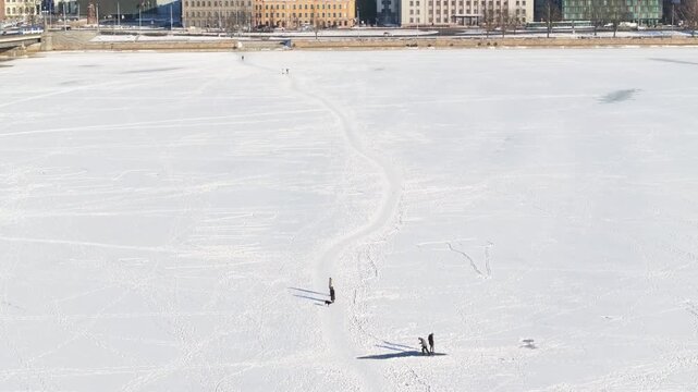 Aerial view of central Riga shows locals and dogs on an S shaped path across frozen Daugava toward the embankment, historic buildings, a bridge left, and long winter shadows.