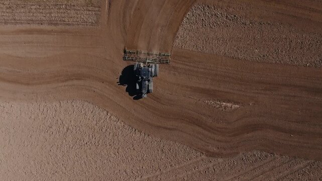 Aerial view shows heavy black tractor pulling cultivator on curved tilled field, dust plume rising, wave like furrows, tire tracks, power pole shadow, daytime