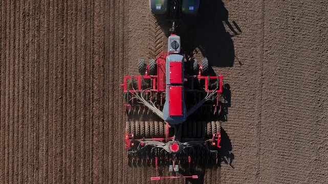 Aerial view shows a red tractor pulling a multi row seeder across brown soil, leaving parallel furrows and tire tracks, with dust from coulters in warm low angle light.