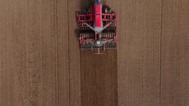 Aerial view of tractor and large multi row seed drill moving down a rectangular field, leaving darker tilled strips, radial hopper pattern, and parallel tire tracks.
