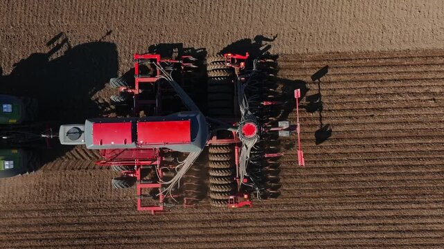 Aerial view shows a green tractor towing a red multi row seeder with disc coulters. Dust lifts as it makes a steady pass, casting long shadows and leaving even furrows.
