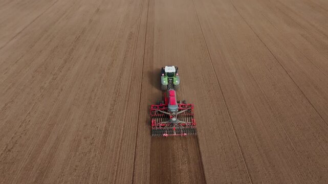 Aerial view shows a tractor towing a seeder across a vast brown field, leaving a darker tilled strip. Parallel furrow lines frame steady forward motion.