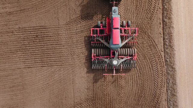 Aerial view shows green tractor pulling red air seeder across tilled brown field. Precision rows form as dusty soil shifts and tire tracks mark steady forward motion.