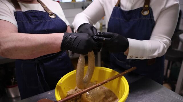 Two people tie sausage links over dowel with yellow bucket and denim. Close up hands in black gloves work with twine and casing in warm light, steady motion.