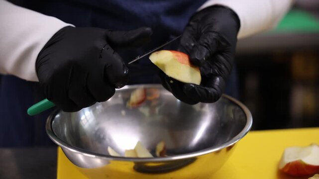 Close view shows black nitrile gloves using green paring knife to peel and slice red skinned apple on yellow board, thin cuts drop into reflective bowl in warm light.
