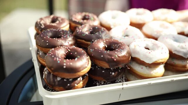 Close view shows stacked chocolate and white glazed donuts with sprinkles on a glass table in bright daylight, slow lateral move and slight rack focus create bokeh.