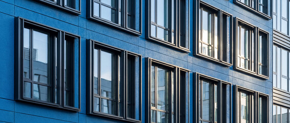 Obraz premium Modern building facade featuring deep blue panels and alternating framed windows reflecting urban environment and light