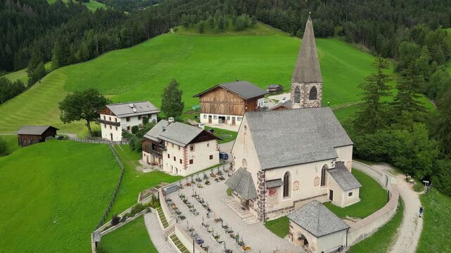 Aerial drone footage circling around the iconic Santa Maddalena church, revealing the cemetery, surrounded by lush green hills in Val di Funes