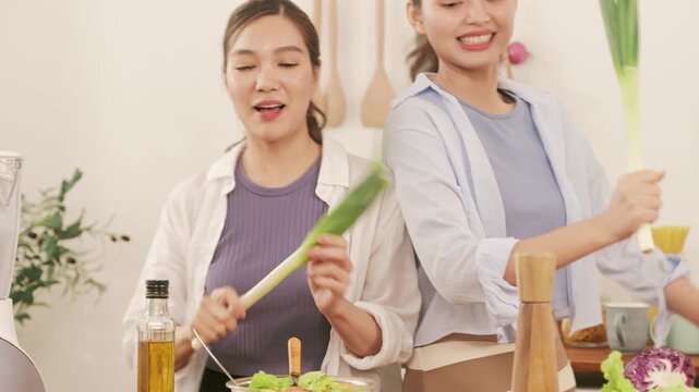 Happy young lesbian couple having fun and dancing with leeks while preparing healthy food in the kitchen.