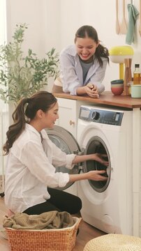 Cheerful Asian lesbian couple doing laundry together at home, loading the washing machine and talking.