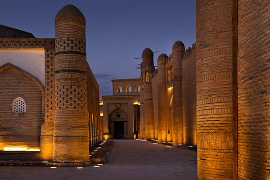Ichan Kala old town illuminated at twilight in Khiva, Uzbekistan. Ancient brick walls and minarets glow with warm ground lights against a deep blue evening sky. Ideal for travel brochures, heritage.