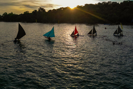 Aerial view of traditional sailboats with colorful sails gliding across the water during a golden sunset in Indonesia.