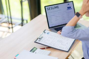 Obraz premium Close up of an Asian female HR manager reviewing and examining job applicant resumes on a clipboard and laptop screen, background checking and recruitment concept.