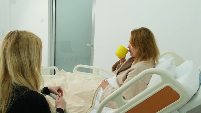Sick woman lies on hospital bed and drinks warm tea as attentive relative sits nearby. Female sips tea during visit from family member. Slow motion