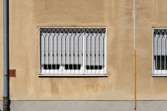 Front view of a rectangular window with white protective iron grilles and closed roller shutters on a textured yellow stucco house wall.
