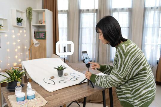 Creative young woman, Online Seller carefully adjusts smartphone and captures professional photos of handmade Jewelry using a simple setup at home