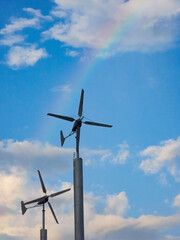 Fototapeta premium Two small wind turbines generate renewable energy against a cloudy blue sky with a colorful rainbow.