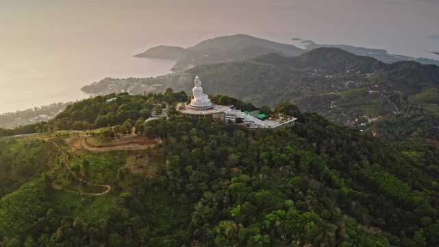 Big Buddha statue sitting on Nakkerd Hill top, overlooking lush green mountains and coastal towns of Phuket, Andaman Sea under soft sunset sky. Thailand travel destination. Aerial view drone panorama