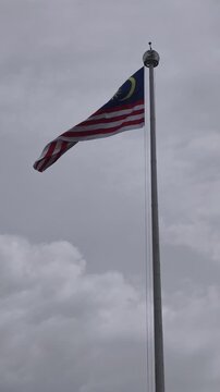 The Jalur Gemilang, national flag of Malaysia, flying proudly on a tall metallic flagpole against a cloudy sky. Featuring fourteen red and white stripes with a blue canton, crescent, and star.