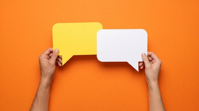 Two human hands holding a bright yellow and a clean white speech bubble cutout made of paper against a vibrant solid orange colored studio surface