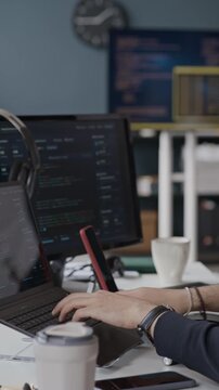 Vertical side view shot of young Middle Eastern male programmer eating takeout lunch at desk and typing on laptop with code on screens. Ideal for tech industry recruiting and software development ads