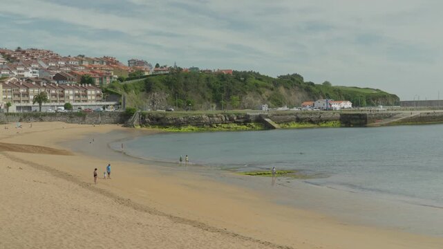 Lovely medium-wide shot of the golden sand beach cove in Luanco, Spain. Features beachgoers along the calm ocean shoreline with traditional coastal apartment blocks sitting on the hills behind.